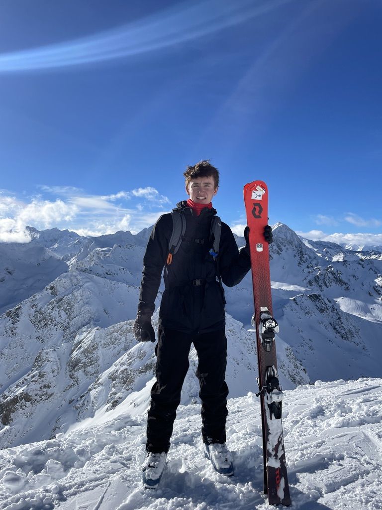 Max Gallinek on a snowy summit holding his skis, with the Alps behind him